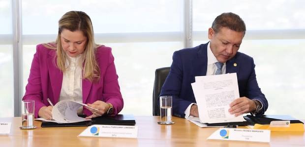 Foto em plano médio de duas pessoas sentadas à mesa, assinando documentos. À esquerda, uma mulhe...