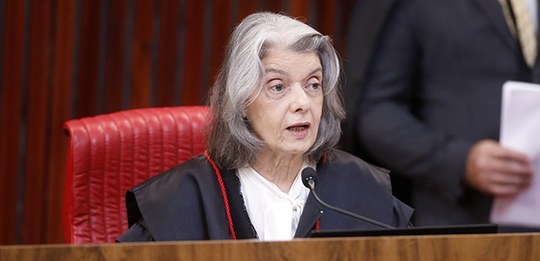 A imagem mostra a ministra Cármen Lúcia, o. Ela veste uma toga preta com detalhes em vermelho. E...